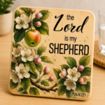 Decorative wooden coaster featuring Psalm 23:1 scripture, "The Lord is my Shepherd," surrounded by beautiful apple blossoms, pink flower buds, green leaves, and a ripe apple. Rustic faith-based coaster with elegant botanical artwork and natural wood finish, perfect for Christian home décor, inspirational gifts, prayer spaces, and religious occasions.